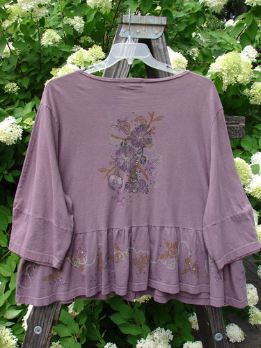 Barclay Peplum Pocket Jacket Magic Garden Dusty Plum Size 2, featuring floral designs, full ceramic button front, and three-quarter sleeves, displayed on a hanger against a wooden structure.