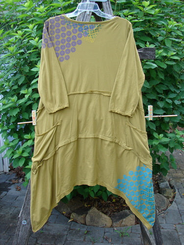 Barclay Raw Seam Vector Tunic Dress in Sectional Daisy Yellow Olive, displayed on a clothesline, showcases raw edges, a curved empire waist, A-line shape, and exterior drop front pockets.