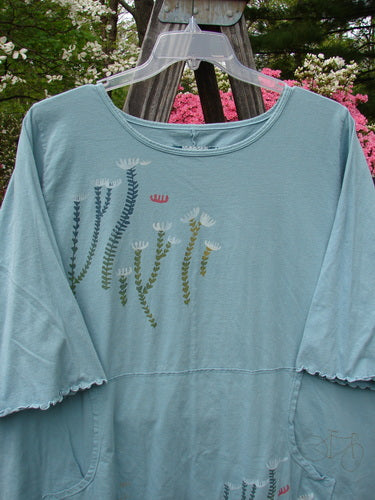 Barclay Three Quarter Sleeved Cafe Pocket Top Fern Blue Tourmaline Size 2, featuring floral patterns, three-quarter sleeves, a rounded hemline, and a kangaroo pocket, displayed on a hanger outdoors.