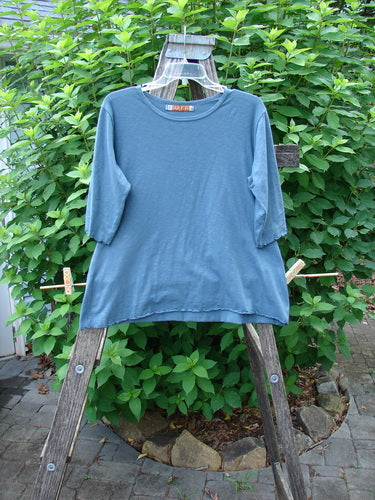 Barclay Slub Three Quarter Sleeved Cafe Tee Unpainted Tealen Size 1 displayed on a clothes rack, showcasing its rounded neckline, slight flare, three-quarter sleeves, and curly edgings in a garden setting.