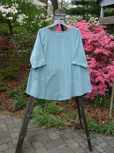 Barclay Three Quarter Sleeved Cafe Pocket Top Fern Blue Tourmaline Size 2 displayed on a wooden hanger, showcasing its longer tunic shape, feminine neckline, and double side entry kangaroo pocket.