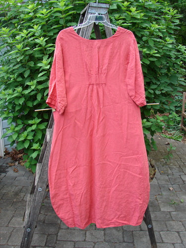 Barclay Linen Ceremony Tea Thostle Dress in Geranium hangs on a clothes rack, showcasing its hourglass shape, upward scoop front hemline, and pleated three-quarter length sleeves, reflecting vintage elegance.