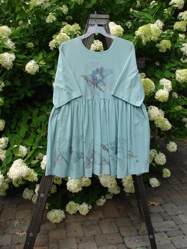 Barclay Flower Garden Tunic Dress Dragonfly Aloe Size 2 displayed on a swing, featuring a rounded neckline, three-button front, open lower front, and a pleated skirt with a hand-painted dragonfly design.