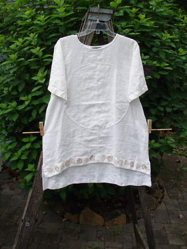 Barclay Linen Shirt Tail Top Circle Center Daisy Chain White Size 0 displayed on a clothes rack, showcasing its rounded neckline, vented hemline, and unique painted circle and daisy border theme.