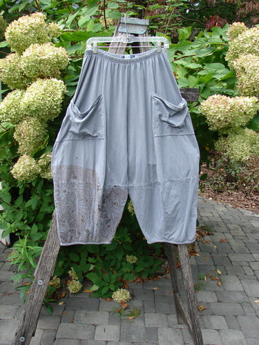 Barclay Crop Two Pocket 4 Square Pant Spin Flower Grey Green Size 0, featuring drop-crotch design, large front pockets, and a distinctive harem style, hangs on a plastic hanger against a wooden frame.