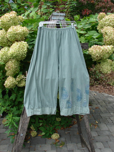 Barclay Flutter Bottom Crop Cargo Pant Medallion Vine Size 0: Loose-fitting, sage-colored cotton pants with a unique flutter bottom, full elastic waist, and dual front floppy cargo pockets, hanging on a wooden ladder.