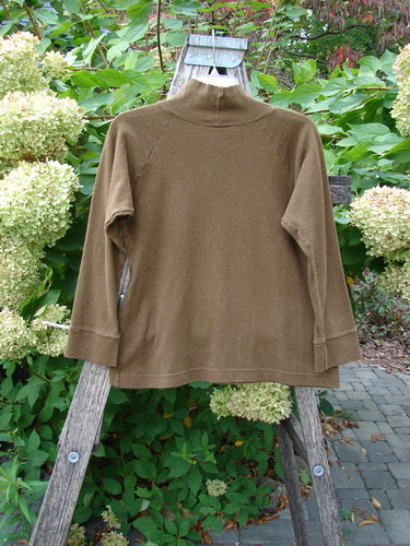 The 2000 Wool Funnel Neck Raglan Top Unpainted Pebble Smaller Size 1 features a mock-neck, long sleeves, and a front cargo pocket, artistically draped on a wooden ladder outdoors.