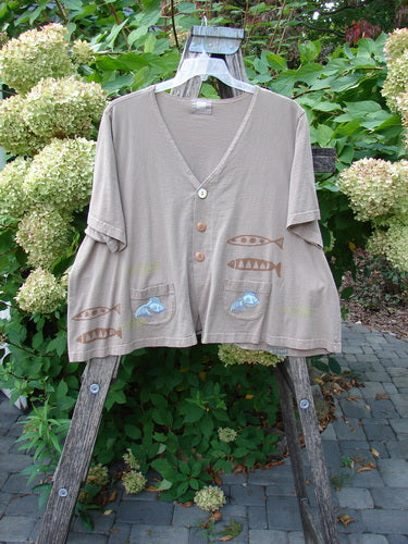 1999 Tiny Pocket Top Fishie Riverbed Size 2: A loose-fitting, earth-toned jacket with fish motifs, V-neck, wide sleeves, and front pockets. Hangs on a wooden ladder outdoors, capturing vintage Blue Fish style.
