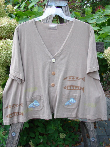 1999 Tiny Pocket Top Fishie Riverbed Size 2: A cropped, loose-fitting cardigan with short sleeves, featuring fish motifs and wooden buttons, displayed on a ladder amidst foliage, embodying vintage Blue Fish Clothing's creative freedom.
