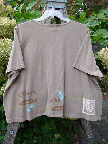 1999 Tiny Pocket Top Fishie Riverbed Size 2 features a taupe loose-fit, short-sleeve T-shirt with a fish-and-wave motif at the hem, displayed outdoors against shrubs and hydrangeas.