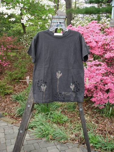 1998 Botanicals Short Sleeved Tee Triple Sprout Raven Size 0 displayed on a wooden rack, showcasing a vibrant flower design and thicker ribbed neckline from the Spring Botanicals Collection.