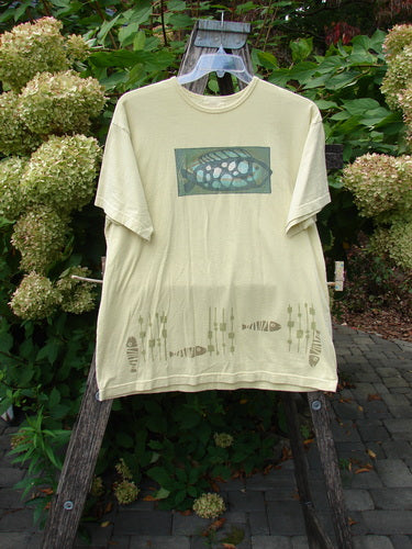 1999 Short Sleeved Tee Single Fish Citron Size 1 displayed outdoors on a wooden ladder, features stylized fish motifs on chest and hem, showcasing a boxed shape and drop shoulders in organic cotton.