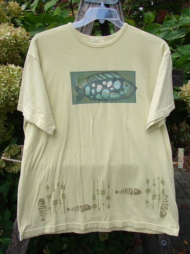 1999 Short Sleeved Tee Single Fish Citron Size 1, featuring an abstract green and white fish design across the chest, with smaller fish and reeds along the hem on organic cotton, hanging outdoors.