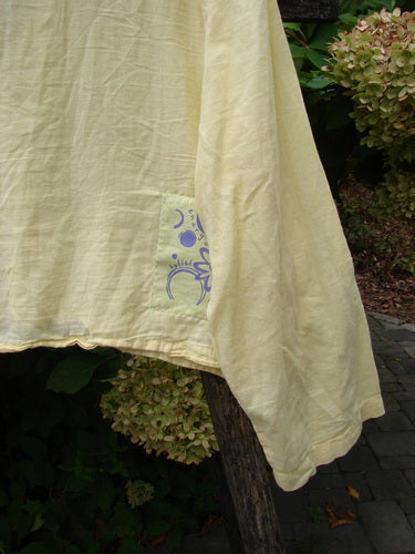 1999 Batiste Boxy Top Unpainted Buttercup Size 2, draped over a wooden post outdoors, features a rounded neckline, purple patch, and billowy sleeves, evoking vintage Blue Fish creativity.