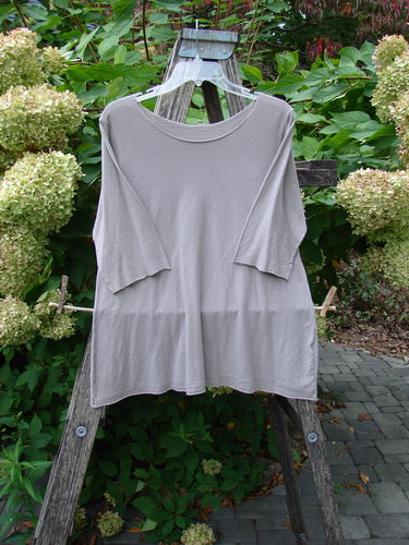Barclay Batiste Raw Edge Layering Top Unpainted Stone Size 1, featuring three-quarter sleeves, raw cut neckline, and rolled edges, hangs on a wooden ladder amidst garden foliage and hydrangeas.