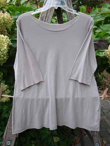 Barclay Batiste Raw Edge Layering Top Unpainted Stone Size 1; a loose-fitting tunic with three-quarter sleeves, raw cut neckline, and rolled edgings, hung on a clear hanger against a wooden ladder.