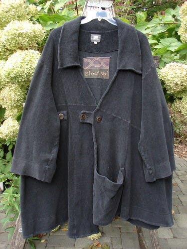 2000 Celtic Moss Highlander Coat Unpainted Black Size 1, featuring a wide draped collar, three-quarter sleeves, oversized pocket, and button tab closure, displayed against hydrangeas, highlighting its vintage Blue Fish charm.