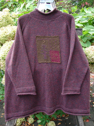 1998 Patched Alpaca Roll Neck Raglan Sweater Wineberry Tweed OSFA features a marled plum color with an olive-green appliqué patch, hand-knitted in a soft wool-blend, displayed against lush garden foliage.