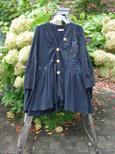 Barclay Thermal Bungalow Jacket Modern Graphics Black OSFA hanging on a ladder, featuring a deep V-neck, wooden buttons, tiered hem, and subtle line-art accents, set in a garden backdrop.