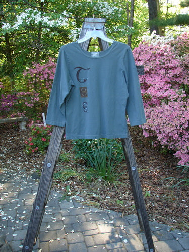 2000 Cotton Lycra Three Quarter Sleeve Layering Top Celtic Park Size 0 displayed on a wooden rack, showcasing its vertical Celtic theme paint and three-quarter sleeves, ideal for layering.