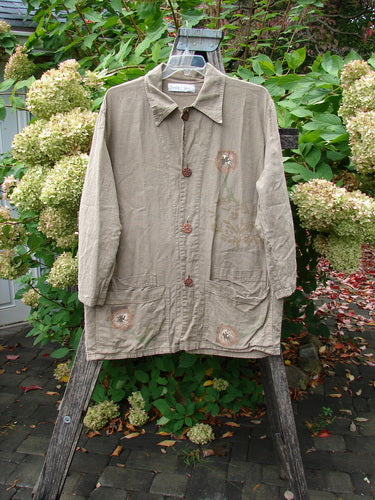 1998 Botanicals Herbary Jacket Dandelion Drift Dust Size 1: light-tan, long-sleeved with wooden-style buttons, subtle floral embroidery, and a pointed collar, hanging on a wooden trellis amidst green foliage and pale hydrangeas.