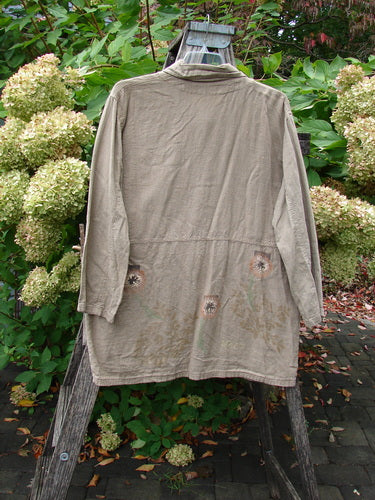 1998 Botanicals Herbary Jacket Dandelion Drift Dust Size 1 draped over a wooden ladder, showcasing its long, khaki design with artistic dandelion-themed paint and ceramic buttons, set in a lush garden.