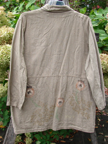 1998 Botanicals Herbary Jacket Dandelion Drift Dust Size 1, showcasing a back view of a muted olive-khaki, long lightweight coat with faded floral motifs on the hem, hanging in a garden setting.