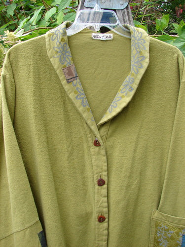 Barclay Patched Flannel Frolic Jacket Green Seed Size 2, featuring an olive-green button-front design with a floral-patterned collar trim, oversized pocket, and unique buttons, hanging outdoors with foliage in the background.