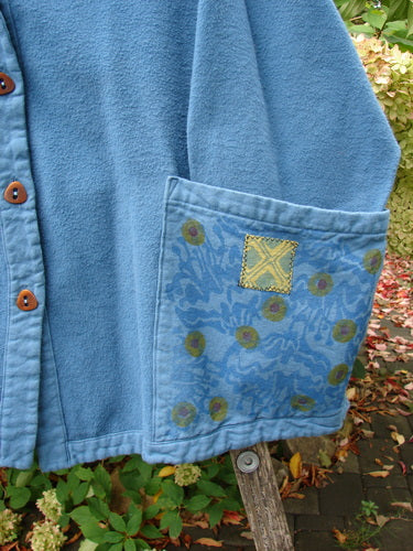 Barclay Patched Flannel Frolic Jacket Garden Blue Teal Size 2, featuring brown buttons, a floral-patterned pocket, and contrasting yellow patch, draped over a chair with garden foliage in the background.