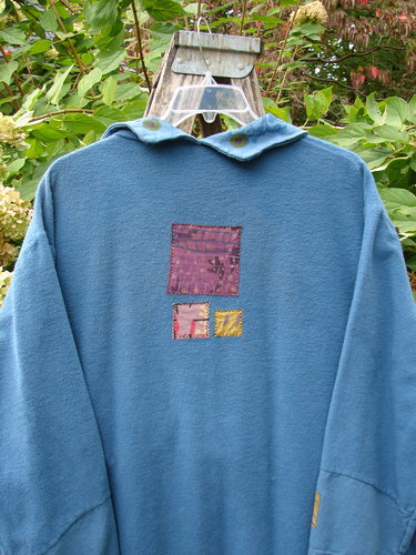 Barclay Patched Flannel Frolic Jacket Garden Blue Teal Size 2 displayed outdoors, featuring three decorative patchwork squares on the back, showcasing its unique design and cozy appeal against lush foliage.