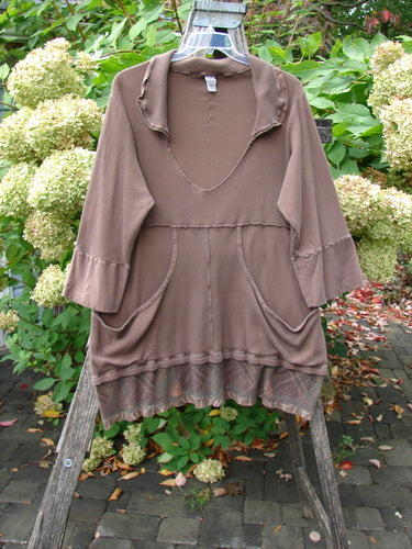Barclay Thermal Flutter Neck Kangaroo Pocket Top Leaf Mud Size 0 features a deep V-neck, ¾-length sleeves, raw-edge seams, and front patch pockets, displayed on a rustic wooden frame in a lush garden.