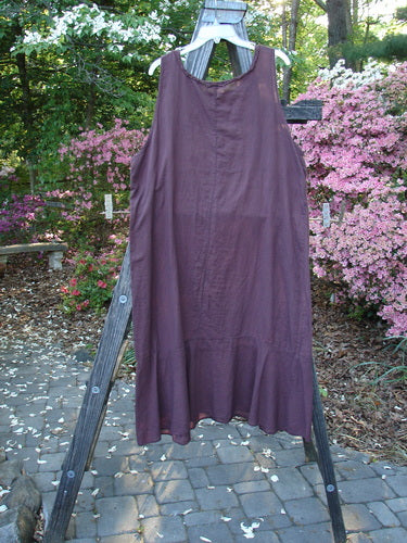 Barclay Batiste Raw Seam Flutter Pinafore Shift Unpainted Fig Size 1 displayed on a wooden rack showcases a ruffled lower, rounded neckline, and downward curved empire waist.