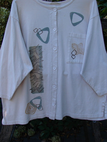 1995 Stargazer Jacket Chair Row China OSFA: A light-cream button-front blouse with abstract geometric prints, featuring a painted oversized pocket, slightly shallow rolled neckline, and original paper buttons, hanging against foliage.