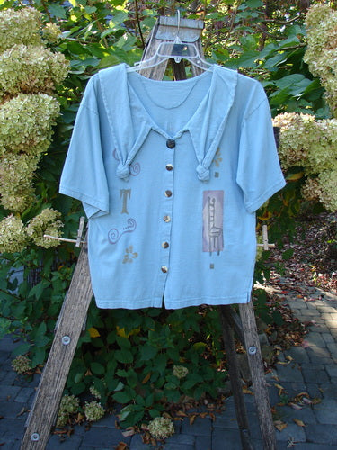 1994 Compass Top Chair Dusk Size 1, light-blue blouse with a scarf-like collar and button-down front, featuring abstract motifs on the lower half, draped on a wooden ladder against a garden backdrop.