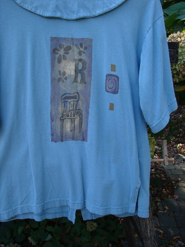 1994 Compass Top Chair Dusk Size 1 features a multicolored graphic with flowers, an R, and an abstract chair. It showcases side vents and a pointed collar, capturing Blue Fish’s vintage charm.