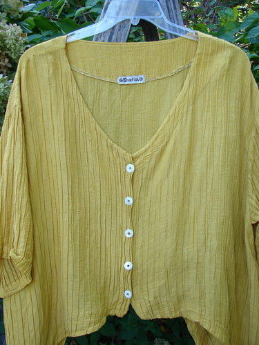 Barclay Rib Silk Dip Side Button Jacket in Golden Sunshine, featuring a V-neck, elbow-length gathered sleeves, and five small white buttons, hanging on a clear plastic hanger outdoors.