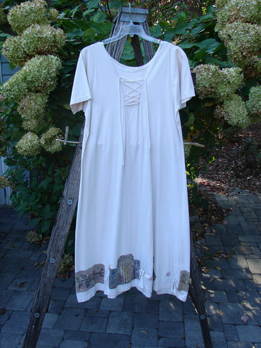 1994 Loop Dress Magic Garden Tea Dye Smaller OSFA hangs on a wooden ladder, showcasing a knee-length, short-sleeved design with lace-up scoop neckline, flared hemline, and decorative hem accents amidst garden greenery.