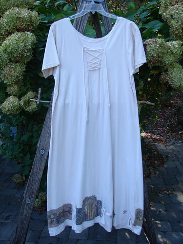 The 1994 Loop Dress Magic Garden Tea Dye Smaller OSFA hangs from a rustic wooden ladder, showcasing its scooped neckline with laced ties and a decorative hem featuring embroidered scenes.