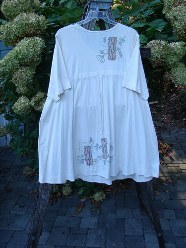 Barclay Tunnel Pocket Pull Over Dress Fish Natural Size 2, featuring elbow-length sleeves, gathered waist, and embroidered motifs, hangs on a wooden ladder outdoors, showcasing its unique style and organic cotton material.