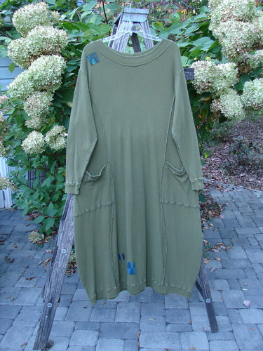 Barclay Patched Thermal Curve Dress Greenroot Size 1 hangs on a wooden ladder outdoors, showcasing its loose fit, patch pockets, raw-edge seams, and unique fabric-patch accents, embodying Bluefishfinder's vintage style.