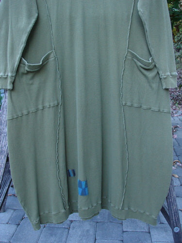 Barclay Patched Thermal Curve Dress Greenroot Size 1 draped over a bench, showcasing its figure-eight shape, exposed seams, front pockets, and a patch accent near the hem.