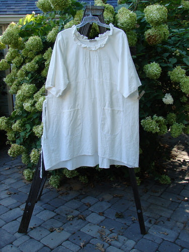 Barclay Linen Duet Sunrise Dress in Crème displayed on a rack, featuring a curly neckline, vertical pleats, pockets, and a varying hemline.