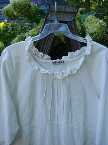 Barclay Linen Duet Sunrise Dress in Crème on a swinger, showcasing a curly cotton neckline, puckered vertical pleats, empire waistline, front pockets, and a varying hemline.