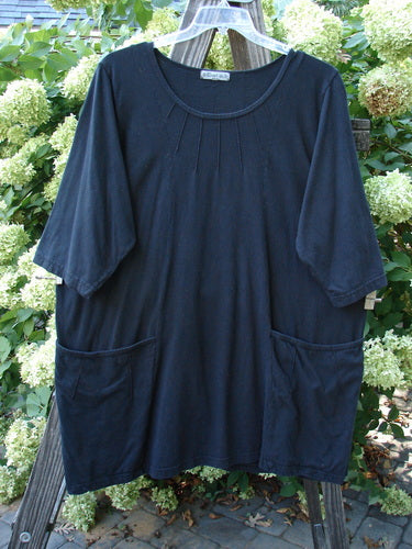 Barclay Sunrise Pin Tuck Pocket Tunic Unpainted Black Size 2 displayed on a hanger, featuring a deeper rounded neckline, A-line shape, short sleeves, and two lower front pockets with pin tucks.