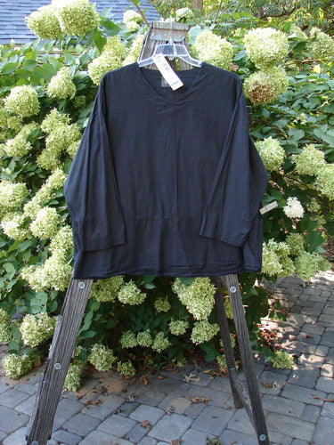 2000 Batiste Overshirt Top Unpainted Black Size 2 displayed on a wooden hanger, featuring a softly rounded V neckline, drop shoulders, and a flowing A-line shape against a floral garden backdrop.
