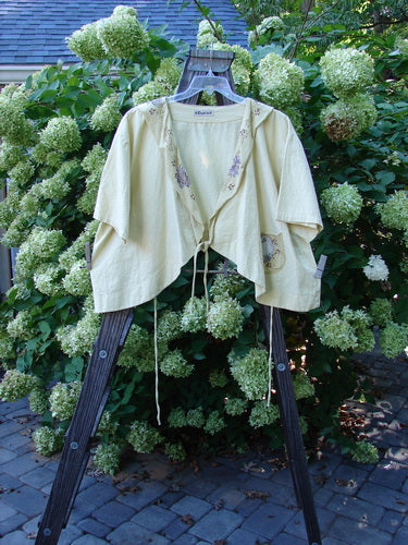 Barclay Linen Batiste Rippie Tie Jacket Shrug with a Morning Floral Theme, displayed on a wooden rack, showcasing a deep V neckline, flutter collar, and varying hemline.