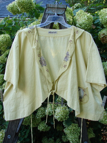 Barclay Linen Batiste Rippie Tie Jacket Shrug Florals Quiet Sunshine Size 2 displayed on a wooden hanger, showcasing its deep V neckline, flutter collar, and painted floral pocket.