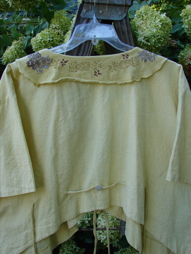 Yellow Barclay Linen Batiste Rippie Tie Jacket Shrug with floral design, featuring deep V neckline, flutter collar, and rear tie back accented by a shell button, displayed on a hanger.