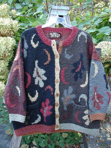 Tara Handknits Star Button Pocket Cardigan Leaf & Moon Brights OSFA draped over a wooden chair in a garden, showcasing its handcrafted patchwork design with abstract motifs and rustic buttons.