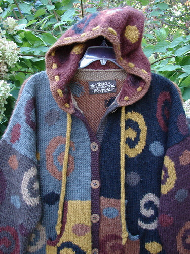 Tara Handknits Hooded Star Button Pocket Cardigan Swirls Brights OSFA features colorful knitted panels with spiral motifs, a pointed hood with draw-cords, oversized star buttons, and a kangaroo pocket, displayed outdoors.